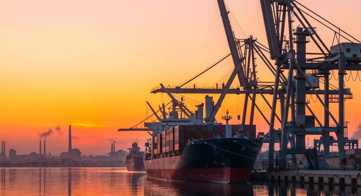 Buques de carga atracados en un puerto industrial al atardecer, rodeados de grúas y reflejados en el agua tranquila