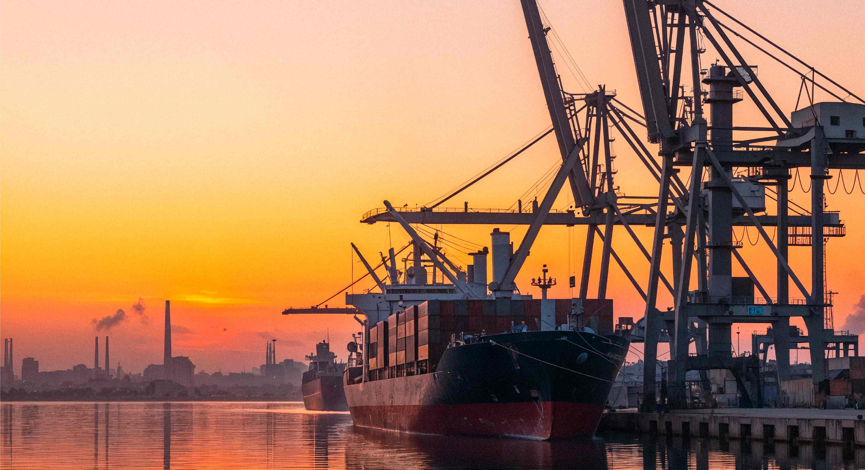 Buques de carga atracados en un puerto industrial al atardecer, rodeados de grúas y reflejados en el agua tranquila