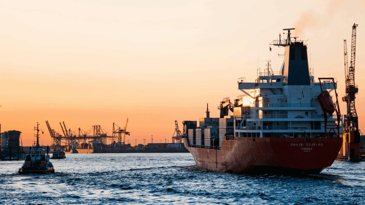 Buque portacontenedores “CHARLES ISLAND” con la palabra “MASAN” navegando en un puerto industrial al atardecer, con grúas y otros barcos al fondo.